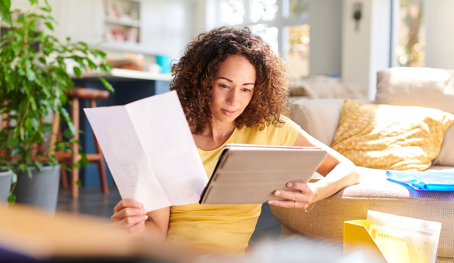 woman in sunny living room holding papers in one hand and her digital tablet in the other