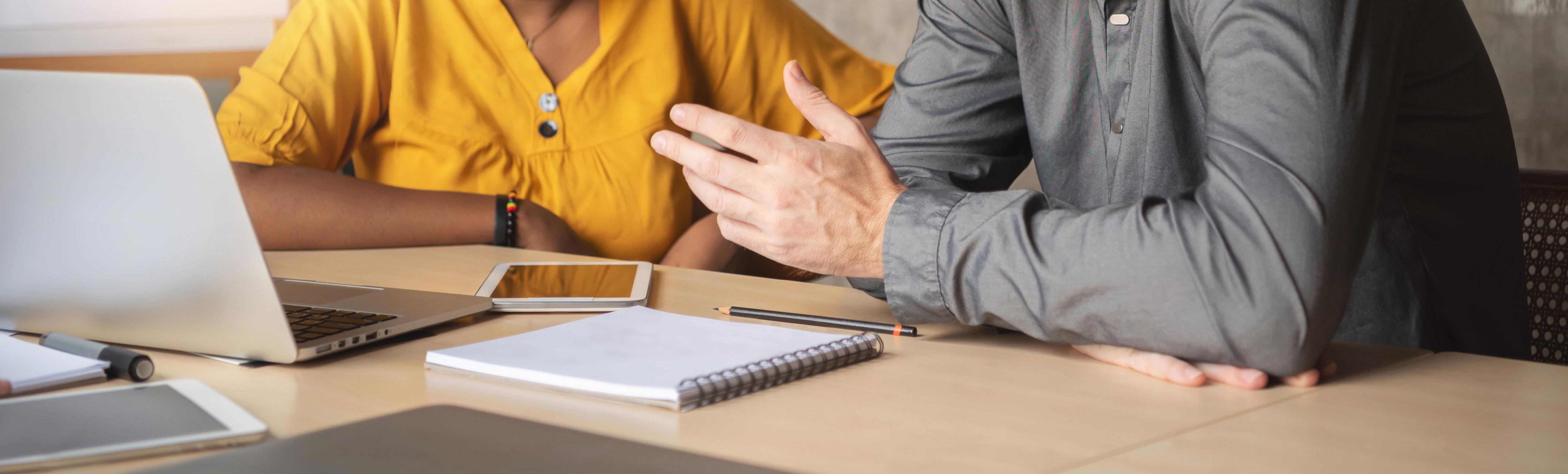 man and woman talking at a desk, cropped between neck and midriff, with gesturing hands